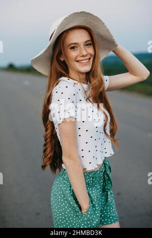 Kaukasische rothaarige Frau mit Hut, die auf einer Straße über einer malerischen Landschaft posiert. Schönes Mädchen. Hübsche junge Frau. Stockfoto