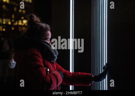 Toronto, Kanada. 07th. Februar 2022. Eine Frau in einem roten Wintermantel nutzt ihre Hand, um mit einer Lichtkunst-Installation ‘Control No Control' von Studio Iregular zu interagieren, einem großen LED-Würfel, der auf alles reagiert, was ihn berührt, und jede Bewegung auf seiner Oberfläche.die Nordic Lights Outdoor-Winterausstellung, Eine Ausstellung nordischer und kanadischer Lichtkunst im Toronto Harbourfront Centre, die gemeinsam mit Fjord Studio produziert wurde. (Foto von Katherine Cheng/SOPA Images/Sipa USA) Quelle: SIPA USA/Alamy Live News Stockfoto