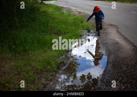 Das Kind spielt neben der Pfütze. Junge zieht Seilspielzeug entlang schlammiger Straße. Kind in orangefarbenem Helm und blauer Jacke geht im Sommer die Straße hinunter. Stockfoto