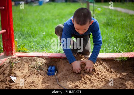 Kind spielt im Sandkasten. Junge in blauer Jacke auf dem Spielplatz. Der Vorschulkinder berührt den Sand mit den Händen. Spaziergang im Sommer im Hof. Kind spielt allein. Stockfoto