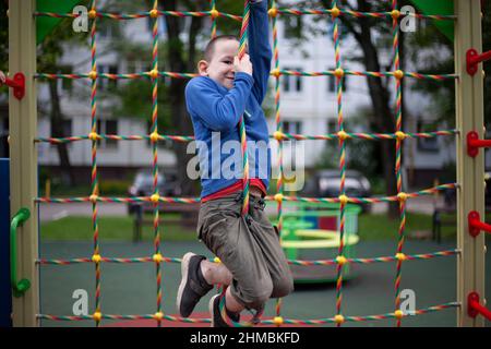 Kind auf dem Spielplatz. Junge klettert eine Seilleiter hoch. Details des Spiels auf der Straße. Kinderbereich im Hof. Geh mit meinem Sohn. Junge hält sich mit seinem h an Seilen fest Stockfoto