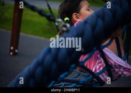 Kind auf runder Schaukel. Kinderspielplatz für Kinder. Asiatische Mädchen schwingt fröhlich. Vorschulkinder spielen im Sommer im Innenhof. Sichere Schaukel für junge ch Stockfoto