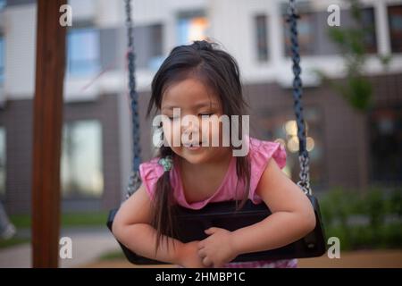 Asiatische Mädchen schwingt auf Schaukel. Kind auf dem Spielplatz. Mädchen spielt im Sommer. Gehen mit Kind auf der Straße. Vorschulkinder genießen Ruhe. Stockfoto