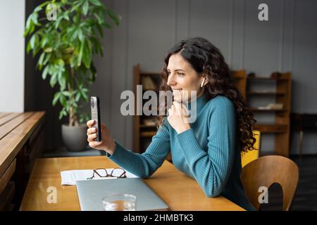 Fröhliche, lächelnde junge Mitarbeiterin in kabellosen Ohrhörern, die das Smartphone für Videoanruf am Arbeitsplatz nutzt Stockfoto
