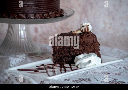 Ein leckeres Stück Schokoladenkuchen wird auf einem schlichten weißen Teller mit einer Schokoladensauce und einer Puppe PF Schlagsahne serviert Stockfoto