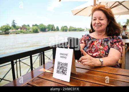 Reife lateinische Frau, die lächelt und einen QR-Code scannt, um auf ein Restaurantmenü zuzugreifen; Verwendung von kontaktloser Technologie im Alltag. Stockfoto