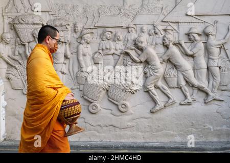 Ein buddhistischer Mönch, der eine Almosenschale trägt, passiert an einem Fries außerhalb der Yodpiman Flower City (Markt), Bangkok, Thailand, und zeigt eine geschäftige Marktszene von früher Stockfoto