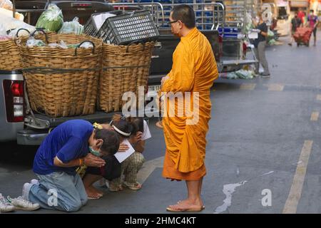 Drei junge Angestellte auf einem Markt in Bangkok, Thailand, knieten tief vor einem buddhistischen Mönch bei seiner morgendlichen Almosenrunde und baten um Segen Stockfoto