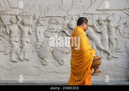 Ein buddhistischer Mönch, der eine Almosenschale trägt, passiert an einem Fries außerhalb der Yodpiman Flower City (Markt), Bangkok, Thailand, und zeigt eine geschäftige Marktszene von früher Stockfoto