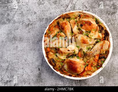 Im Ofen gebackenes Huhn und Reis mit Zwiebeln, Karotten und Kräutern in einer weißen Backform auf dunkelgrauem Hintergrund. Draufsicht, flach liegend Stockfoto