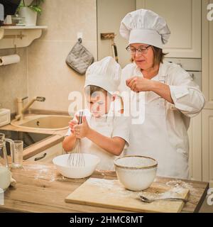 Mutter und Sohn kochen Apfelkuchen in der heimischen Küche. Eine Frau und ein Junge in Küchenhüten und Schürzen kochen mit Gebäck Stockfoto