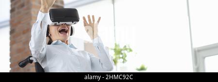 Frau, die im Rollstuhl sitzt und eine Virtual-Reality-Brille trägt und ihre Hände winkt Stockfoto