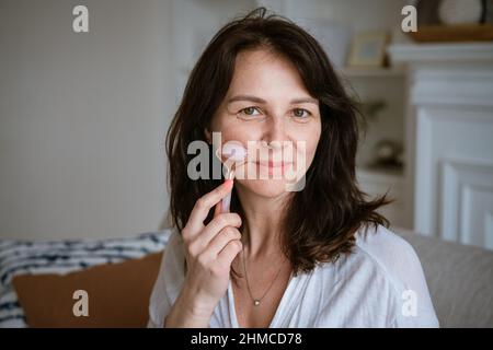 Porträt einer kaukasischen Frau, die Gesichtsmassage mit Quarzschaber für rosa Gouache macht, Robe trägt, die auf dem Sofa im gemütlichen Wohnzimmer zu Hause sitzt, Anti-Aging-Schönheitsroutine-Konzept Stockfoto