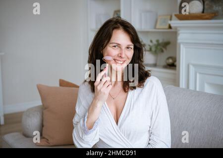 Gesichtsmassage. Portrait nette gepflegte Frau im Bademantel auf der Couch zu Hause. Brünett kaukasische Frau mit gesunder, gepflegter Haut. Stil und Schönheit, Hautpflegekonzept Alternative Gesichtsbehandlung Stockfoto