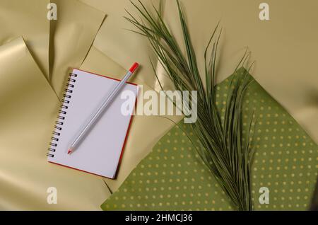 Ein offenes leeres Notizbuch und ein Bleistift. Ein Palmenkranz und ein grünes Stück Papier vor beigem Hintergrund. Abgewinkelte Ansicht von oben. Selektiver Fokus Stockfoto