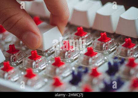 Perspektivische Ansicht Nahaufnahme von Kinderfingern, die eine benutzerdefinierte Tastenkappe auf einer weißen mechanischen Gaming-Tastatur mit roten Kirschholzschaltern schalten Stockfoto