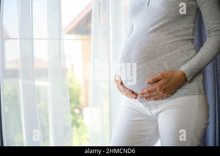 Schwanger Frau mit den Händen auf dem Bauch stehen in der Nähe des Fensters, Mutterschaft Konzept Stockfoto