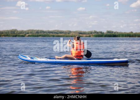 Blonde kleine Mädchen sup Boarding auf See bei Tag Rudern mit Ruder mit Schilf und Bäume im Hintergrund tragen Weste Schwimmweste. Aktivurlaub Stockfoto