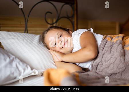 Lächelndes junges kaukasisches Mädchen, das die Kamera auf einem weichen Kissen auf einem großen Doppelbett sieht, wobei die Hände unter dem Kopf mit einer warmen Decke bedeckt sind. Aufwachen Stockfoto