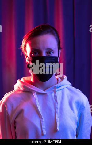 Eine junge, schöne Frau in einem weißen Kapuzenpullover zieht während der Coronavirus-Pandemie eine schwarze medizinische Maske auf einen blau-violetten Hintergrund. Selektiv Stockfoto