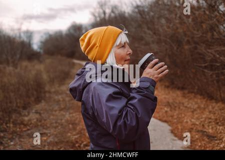 Ältere Frau, die im Herbstpark der Stadt Kaffee zum Mitnehmen hält. Stockfoto