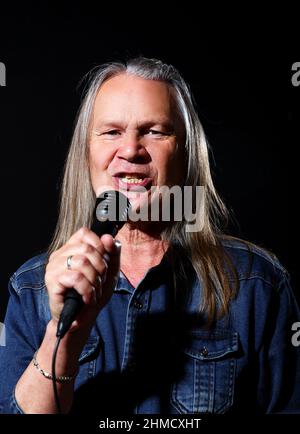 Älterer Mann mit langen grauen Haaren in blauem Denim-Hemd singt auf schwarzem Hintergrund in ein Mikrofon auf der Bühne Stockfoto