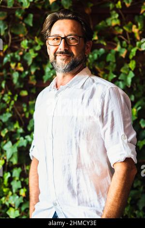 Porträt eines erwachsenen, hübschen bärtigen Mannes mit weißem Hemd, der die Kamera anlächelt und eine Brille trägt. Laub Blatt grün Hintergrund Vertikal komposit Stockfoto