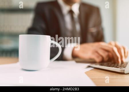 Kaffeebecher-Mockup-Bild, weißer Becher auf Büroschreibtisch mit Geschäftsmann, der im Hintergrund am Laptop-Computer arbeitet, selektiver Fokus Stockfoto