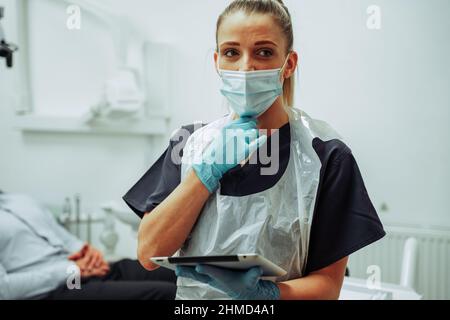 Kaukasische Krankenschwester, die eine OP-Maske trägt und eine digitale Tablette hält, bevor sie den männlichen Patienten beurteilt Stockfoto