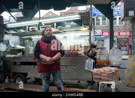 Eine südamerikanische Frau, die gegrilltes Fleisch und andere Lebensmittel aus einem Wagen direkt an der Roosevelt Ave. Und unter den erhöhten U-Bahn-Zügen verkauft. Stockfoto