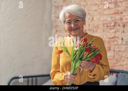 Oma's Day Muttertag Ältere kaukasische Frau in einem gelben Hemd grinsend und hält rote Tulpen mittlere Aufnahme . Hochwertige Fotos Stockfoto