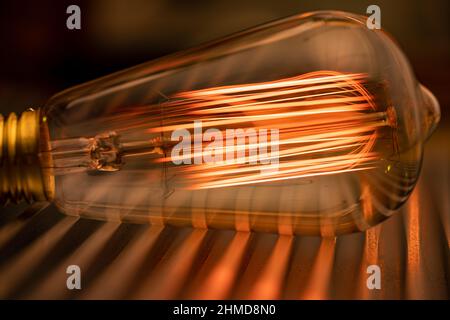 Eine helle, transparente Glühlampe aus Glas leuchtet mit einem schwachen, bernsteinfarbenen, feurigen Licht, das auf einer glatten Wellmetalloberfläche in der Werkstatt eines Ingenieurs liegt Stockfoto