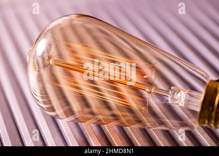 Eine helle, transparente Glühlampe aus Glas leuchtet mit einem schwachen, bernsteinfarbenen, feurigen Licht, das auf einer glatten Wellmetalloberfläche in der Werkstatt eines Ingenieurs liegt Stockfoto