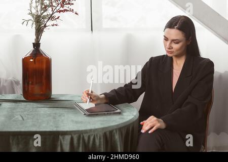 Junge Brünette Freiberuflerin Designerin zeichnet mit einem Stift Skizzen auf einem Grafiktablett. In einem eleganten schwarzen Anzug gekleidet. Klassisches Interieur Stockfoto