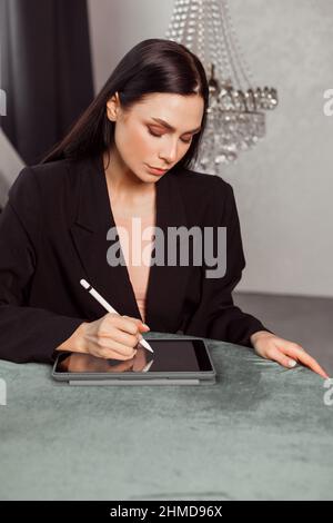 Junge Brünette Freiberuflerin Designerin zeichnet mit einem Stift Skizzen auf einem Grafiktablett. In einem eleganten schwarzen Anzug gekleidet. Klassisches Interieur Stockfoto