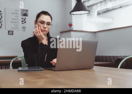 Junge Brünette Freiberuflerin Frau in schwarzem Anzug procrastinate mit Laptop trinken Kaffee in leerem Café. Verschwenden Sie Zeit im Internet Stockfoto