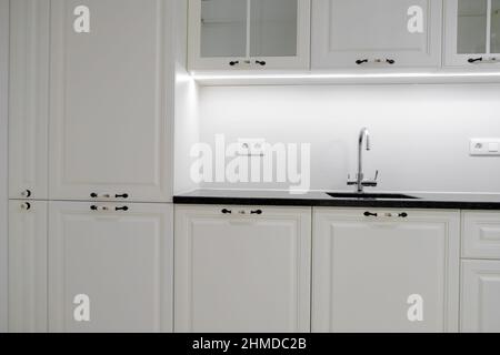 Moderne und elegante Küche mit weißen Schubladen und Schränken, Arbeitsplatte aus schwarzem Marmor und eingebautem Waschbecken mit silbernem Wasserhahn Stockfoto
