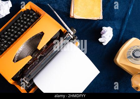Draufsicht auf Vintage-Schreibmaschine mit leerem Papier, Telefon und altem Buch auf blauer Tischdecke Stockfoto