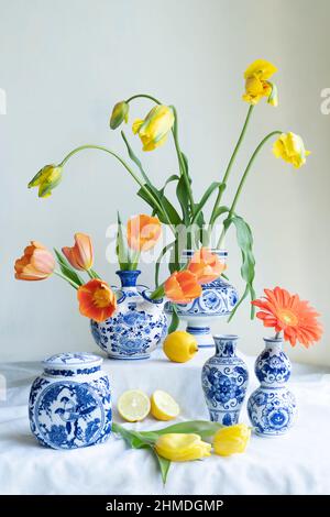 Üppiges natürliches Licht stillife mit alter holländischer Delfter blau lidierter Vase mit gelben und orangen Tulpen, Gerbera und Zitronen auf weißem Leinen gegen ein weißes b Stockfoto