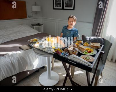 Junge sitzt auf einem Stuhl neben einem frisch zubereiteten Frühstück, das auf dem Tisch im Hotelzimmer serviert wird Stockfoto