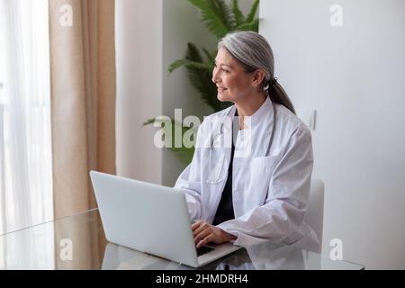 Porträt einer glücklichen reifen Ärztin, die im Büro am Laptop arbeitet Stockfoto