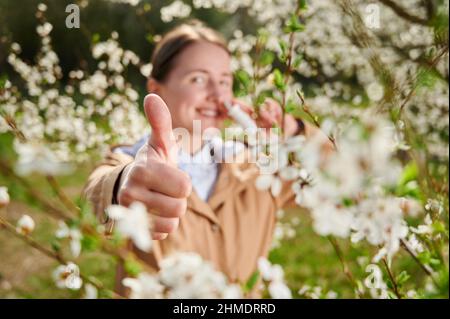 Allergische Frau, die im Frühling an einer saisonalen Allergie leidet und im Frühling im blühenden Garten posiert. Glückliche Frau mit nasalen Tropfen, zeigt Daumen nach oben. Konzentrieren Sie sich auf eine Hand. Frühjahrsallergiekonzept Stockfoto