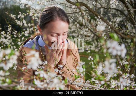 Allergische Frau, die im Frühling an einer saisonalen Allergie leidet und im Frühling im blühenden Garten posiert. Junge Frau niest unter blühenden Bäumen. Frühjahrsallergiekonzept Stockfoto
