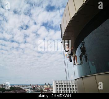 Industriearbeiter beim Bergsteigen, der am Kletterseil hängt und Glasfenster des Wolkenkratzers putzt. Männlicher Reiniger, der beim Waschen der Fenster eines Hochhauses Sicherheitsausrüstung verwendet. Stockfoto