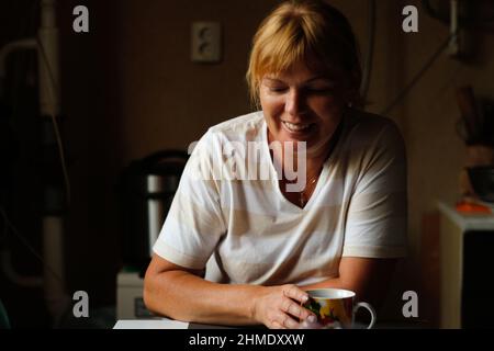 Unschärfe glücklich junge kaukasische Frau lächeln, während zu Hause mit Kaffee oder Tee Tasse sitzen. Morgendliche Routine in der Küche. Lifestyle zu Hause. Frohe 40 Jahre w Stockfoto