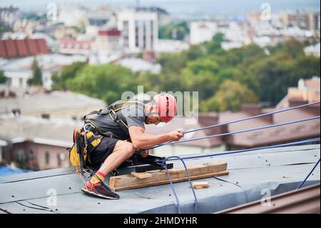 Industriebergsteiger Arbeiter in Schutzhelm mit Sicherheit Kletterseil beim Klettern Dach. Mann, der während der industriellen Hochhausarbeiten Sicherheitskletterausrüstung verwendet. Stockfoto