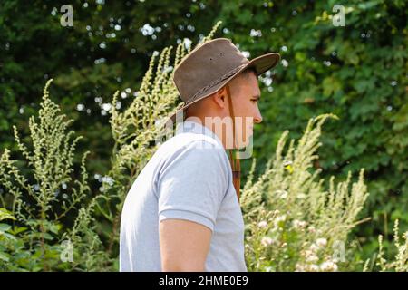 Rüde im Hut. Porträt eines lächelnden Bauern mit grünem Gras und Bäumen Natur im Hintergrund. Junger Mann mit Cowboyhut auf dem Feld. Nahaufnahme. Frohes junges p Stockfoto