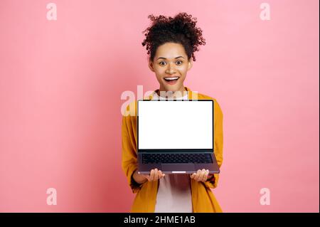 Aufgeregt dunkelhäutig positive junge Frau in Freizeitkleidung, mit offenem Laptop mit leerem weißen Bildschirm, steht auf isoliertem rosa Hintergrund, schaut auf die Kamera, mockup für Ihre Werbung, Kopierraum Stockfoto