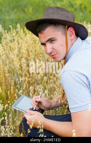 Digitale Farm. Porträt eines Bauern, der im goldenen Weizenfeld sitzt und auf dem Tablet scrollt. Junger Mann mit Cowboyhut auf dem Feld, der Weizenernte untersucht. Hafer Stockfoto