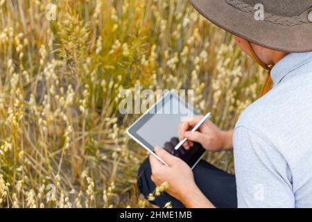 Digitale Farm. Porträt eines Bauern, der in einem goldenen Weizenfeld sitzt und auf einem Tablet schreibt. Junger Mann mit Cowboyhut auf dem Feld, der Weizenernte untersucht. Hafer g Stockfoto
